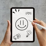 Hands holding a tablet with hand-drawn doodles including a smiley face, heart, megaphone, and light bulb icons