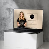 Laptop on white pedestal displaying woman in black shirt and beauty brand logo on beige background
